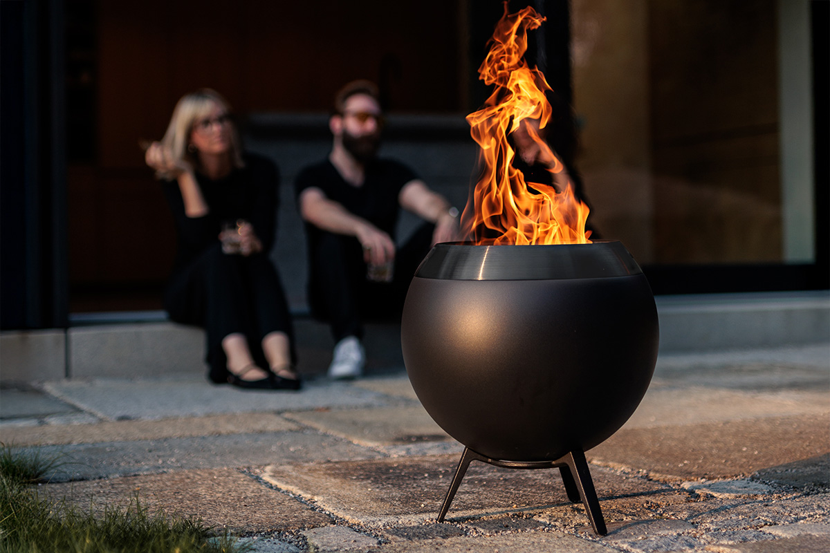 MOON 45 all black Fire Basket with low stand Im Vordergrund ist MOON mit niedrigem Fuß zu sehen im Hintergrund zwei schwarz gekleidete Personen, die ins Feuer schauen.