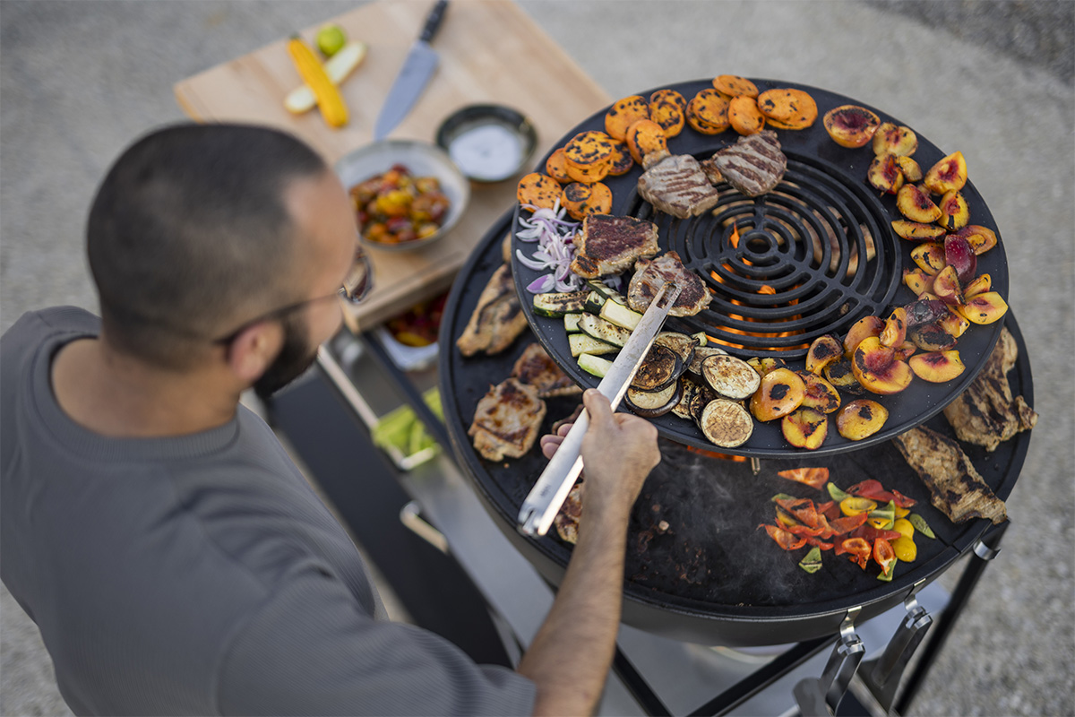 BOWL 70 Anneau Chauffant Le 00617 BOWL 70 Warming Ring est présenté alors qu'un homme Grille sur un grand gril avec une pince, créant une image élégante et accessible.