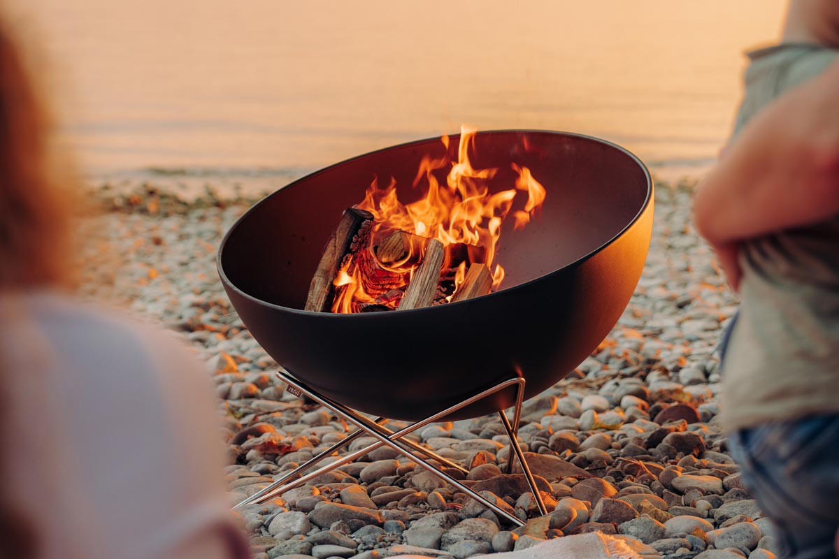 Une femme assise devant un brasero, avec le bol 00249 BOWL 57 avec support en étoile ajoutant une touche fonctionnelle et élégante à la scène.
