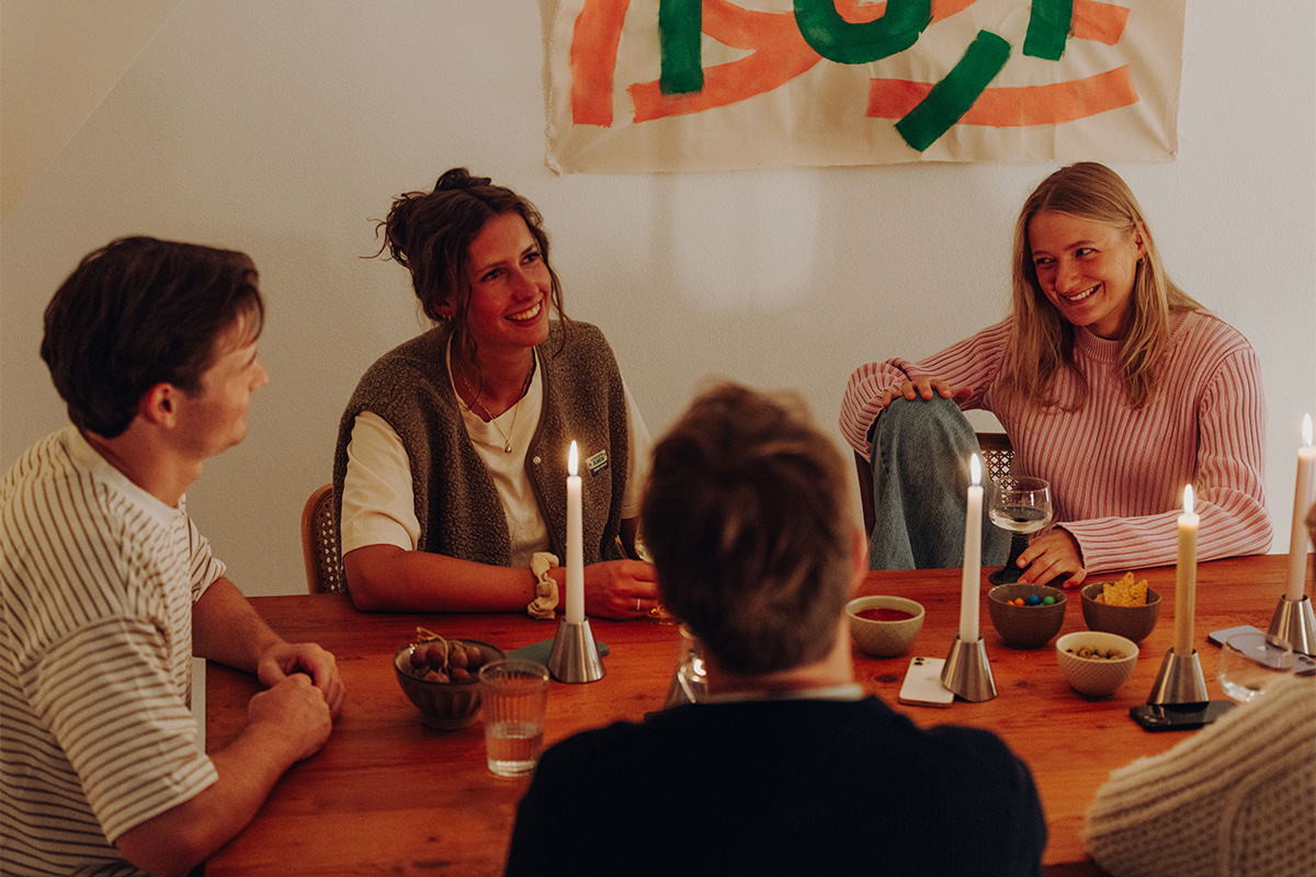 Fünf Personen sitzen ins Gespräch vertieft an Esstisch. Dabei liegen die Smartphones jeweils unter einem Digi Detox Kerzenhalter.